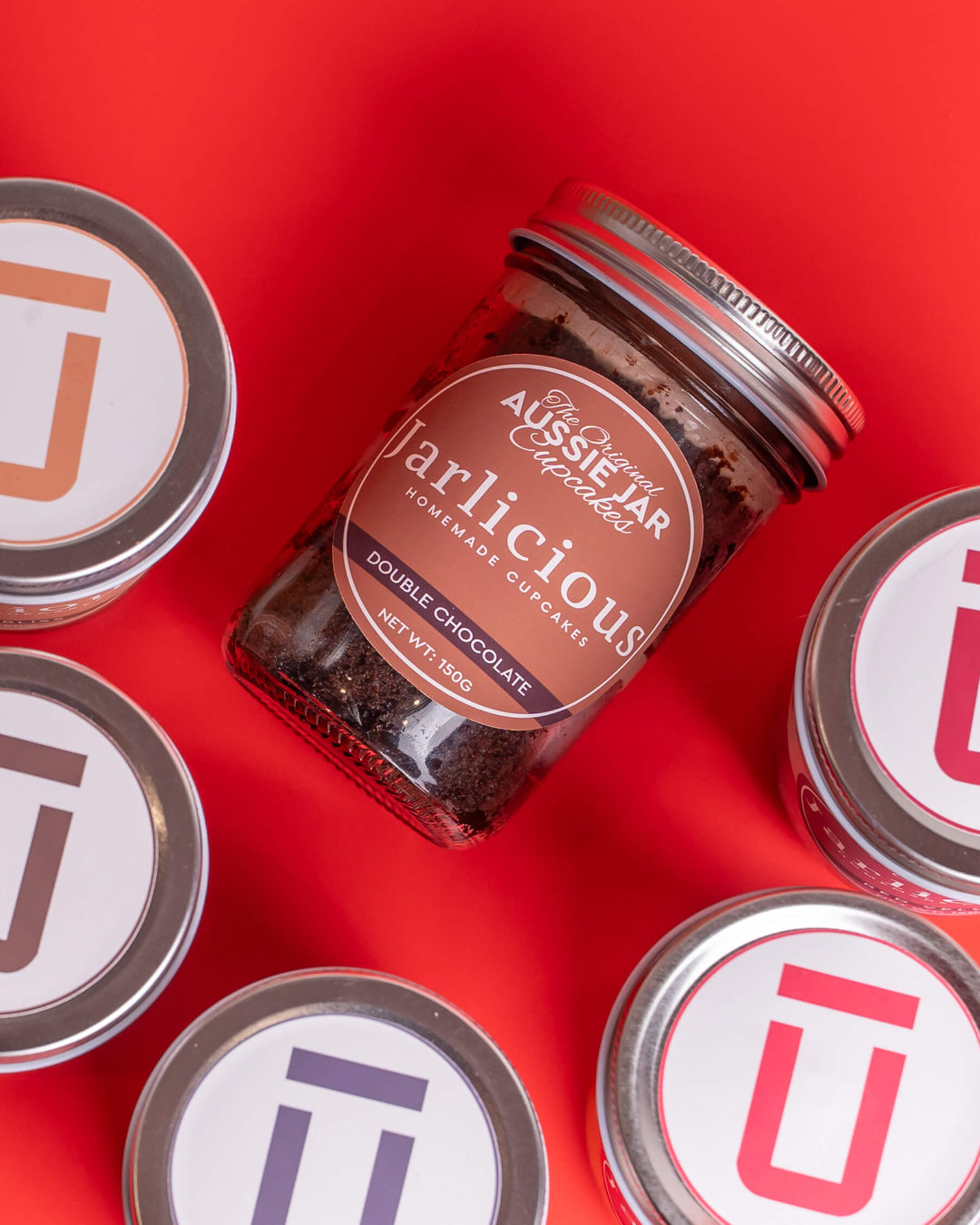 A glass jar of Aussie Jar 'Jarlicious' double chocolate gourmet cupcakes arranged on a bright red background, surrounded by several other jars with white lids featuring colorful Jarlicious Cupcakes logos.